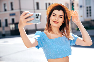 Wall Mural - Young beautiful smiling hipster woman in trendy summer blue clothes. Sexy carefree woman posing on the street background at sunset. Positive model outdoors. Cheerful and happy and hat, take selfie