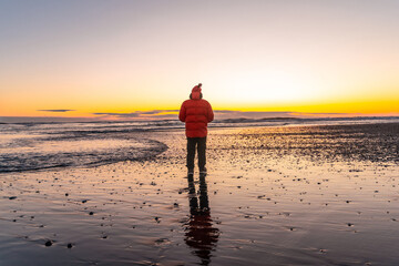 A man stands on a beach at sunset, wearing a red jacket