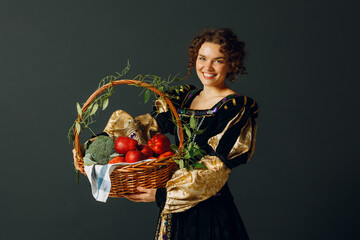 Wall Mural - Portrait of a young adult woman dressed in a medieval dress holding a basket with vegetables and fruits, apples and brussels sprouts. Harvest concept.