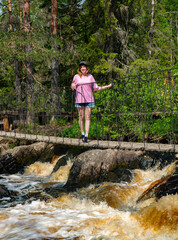 Wall Mural - A woman in a pink shirt and denim shorts stands on a suspension bridge over a rushing river in a forest. The lush greenery and the sound of the flowing water create a refreshing outdoor scene, perfect