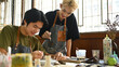 © saltdium - An art teacher assists a student in sculpting clay at a pottery wheel. Both are focused and smiling in a bright studio filled with art tools, paint supplies, and natural light from large windows