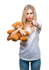 Sticker - Young beautiful blonde woman holding teddy bear over isolated background pointing with finger to the camera and to you, hand sign, positive and confident gesture from the front