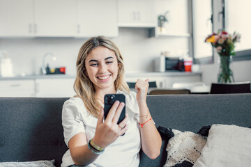 Positive cheerful young woman looking at cellphone screen, getting message with amazing news, celebrating online lottery auction betting win, feeling overjoyed at home, internet success concept..