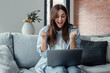 © Daniel - Excited emotional screams with joy woman sit on sofa hold on lap notebook makes Yes hand gesture feels happy getting job of dream, e-mail sales discount special offer, on-line lottery victory concept.