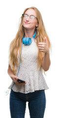 Poster - Blonde teenager woman holding smartphone and wearing headphones happy with big smile doing ok sign, thumb up with fingers, excellent sign