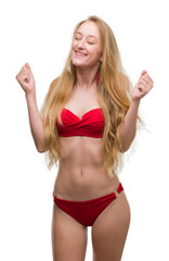 Wall Mural - Blonde teenager woman wearing red bikini excited for success with arms raised celebrating victory smiling. Winner concept.