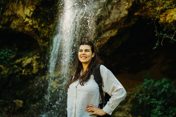 One young caucasian woman is taking a break from hiking near the river and waterfall in the forest	