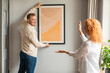 © Home-stock - Young spouses arranging new flat, hanging up painting on wall. New home and relocation concept
