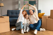 © Home-stock - Happy young parents and kid promoting mortgage, real estate property buying, sitting on floor in new apartment with boxes behind, making hand roof gesture