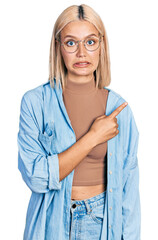 Sticker - Beautiful young blonde woman wearing glasses pointing aside worried and nervous with forefinger, concerned and surprised expression