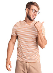 Wall Mural - Young caucasian man wearing casual clothes and glasses smiling with happy face looking and pointing to the side with thumb up.