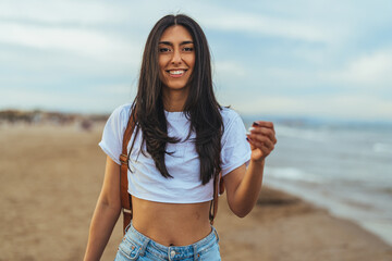 A young Hispanic woman smiles brightly, wearing casual beach attire and standing by the shore, embodying the essence of a relaxed coastal lifestyle.