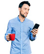 © Krakenimages.com - Hispanic man with beard using smartphone and drinking a cup of coffee smiling looking to the side and staring away thinking.