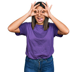 Poster - Young hispanic girl wearing casual purple t shirt doing ok gesture like binoculars sticking tongue out, eyes looking through fingers. crazy expression.