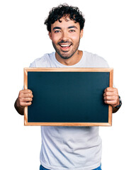 Canvas Print - Hispanic young man with beard holding blackboard smiling and laughing hard out loud because funny crazy joke.