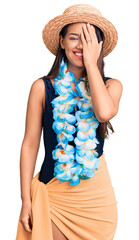 Wall Mural - Young beautiful latin girl wearing hawaiian lei and summer hat covering one eye with hand, confident smile on face and surprise emotion.