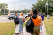 © Alberto - Young woman with backpack and headphones walks towards a group of classmates as they arrive at the university camp or high school after vacation.