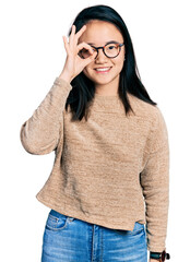 Wall Mural - Young chinese woman wearing casual sweater and glasses doing ok gesture with hand smiling, eye looking through fingers with happy face.