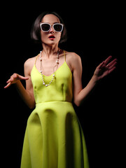 stylish brunette with short hair in white sunglasses in a yellow dress on a black background artistic light