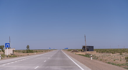 Wall Mural - A long, empty road with a blue sign on the side. The sky is clear and blue