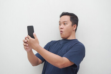 Young Asian man looking smartphone with shocked and surprised expression.  Male with blue t-shirt showing excited expression isolated on white background