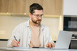 © Ivan - Happy handsome man with laptop and note pad at the kitchen table work remotely from home