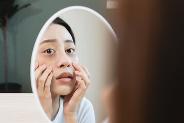 Sleepless Asian woman looking her face in the mirror and worry about dark circles under eyes