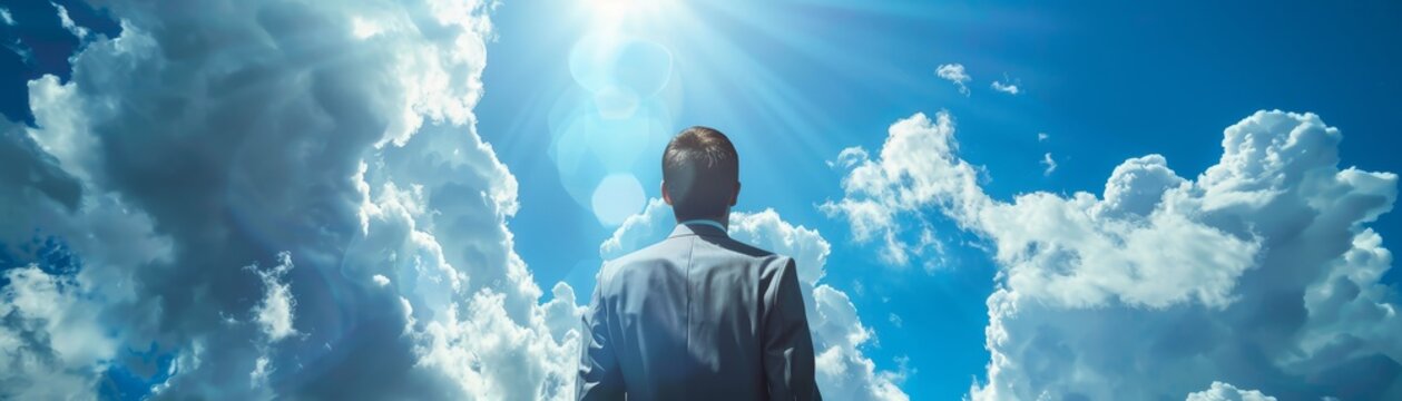 A man in a suit stands facing away from the camera, looking up at a bright sky with clouds.