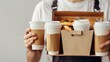 © Emil - Holding various take-out food containers, pizza box, coffee cups in holder and paper bag, close-up. Light grey background, place to insert your text. Delivery man.