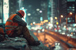 © KirKam - Weary construction worker taking a break at a city construction site during the night, surrounded by urban lights.