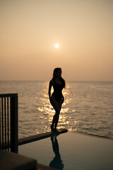 Wall Mural - Woman in black swimsuit relaxing by infinity pool with ocean view during sunset.