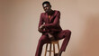 © Generative AI - full length of young african american model in burgundy suit sitting on wooden high stool on beige : Generative AI