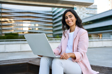 Wall Mural - Smiling young middle eastern Israel businesswoman using laptop pc, looking at camera portrait. Indian or arabic woman freelancer in suit doing business analytics on computer, working remotely outdoors