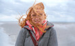 © ullision - A woman with red hair stands on a windy beach, her hair blowing wildly. She wears a gray jacket and pink hoodie, smiling despite the blustery weather, background shows a cloudy overcast sky
