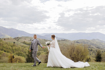 Wall Mural - A bride and groom are walking on a grassy hillside, with the bride wearing a long white dress. The scene is serene and picturesque