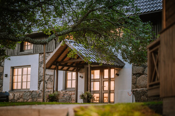 Blome, Latvia - September 11, 2023 -  A cozy rustic house entrance with warm lights glowing from windows, surrounded by stone and wood elements, partially obscured by tree branches.