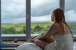 © ullision - red-haired woman sits on bed gazing out large window. lush green landscape under rainy cloudy sky, white lace dress, she exudes a peaceful contemplative mood reflecting on the serene view