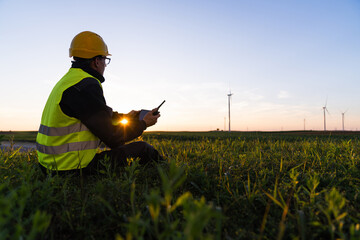 Aufkleber - Worker with digital tablet sits on the grass and looks at wind turbines.