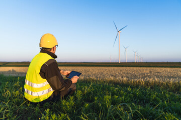 Aufkleber - Worker with digital tablet sits on the grass and looks at wind turbines.