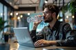 © spyrakot - Young Professional Drinking Water While Working on Laptop in Modern Office - Promoting Productivity and Health