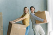 © Ilona - Happy adult women relocating house lifting cardboard box with life stuffs. Friend helping.