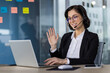 © Liubomir - Businesswoman wearing a headset and waving during a video conference call while sitting in a modern office environment.