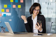 © Liubomir - Happy businesswoman celebrating success while reading good news on her smartphone in office, sitting at a desk with a laptop.