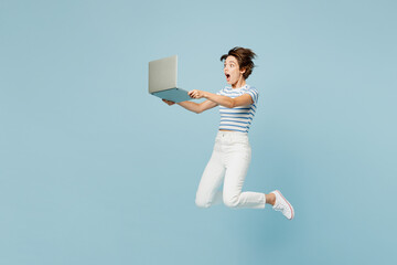 Wall Mural - Full body side view young surprised shocked IT woman wears striped t-shirt casual clothes jump high hold use work on laptop pc computer isolated on plain light blue cyan background. Lifestyle concept.