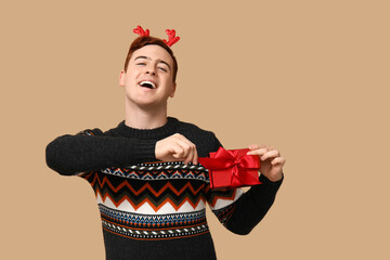 Sticker - Handsome young man in warm sweater and reindeer headband with gift box on beige background