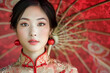 © Cadengo - A beautiful Asian woman elegantly poses while wearing a traditional Chinese dress, complemented by a red umbrella
