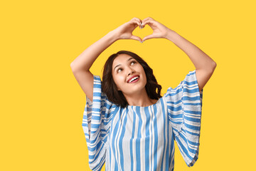 Wall Mural - Beautiful young Asian woman showing heart gesture on yellow background. Valentine's Day celebration
