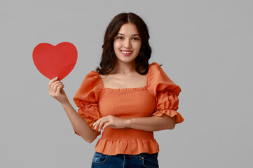 Wall Mural - Happy young Asian woman with red paper heart on grey background. Valentine's Day celebration