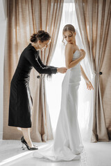 Wall Mural - A woman in a wedding dress is getting her dress altered by a woman in a black dress. The scene is set in a room with curtains