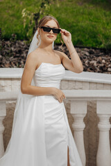 Wall Mural - A woman in a white dress is wearing sunglasses and holding her hands in her pockets. She is standing in front of a white railing